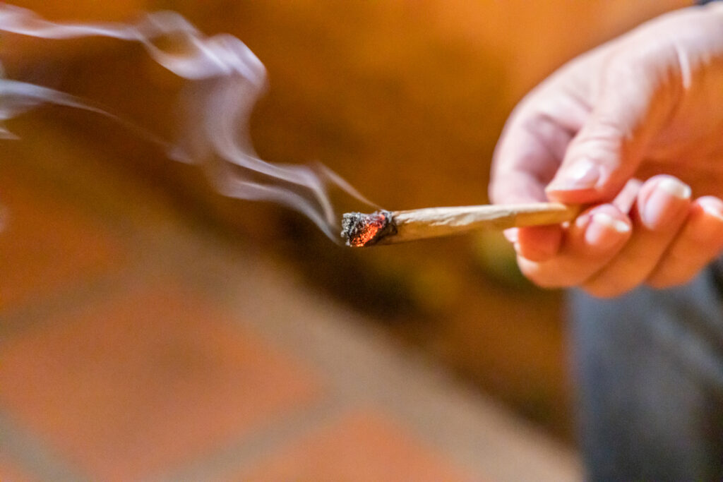 Finger holding lighted cannabis cigarette with smoke in dark room, with selective focus on cannibis ash and fire.